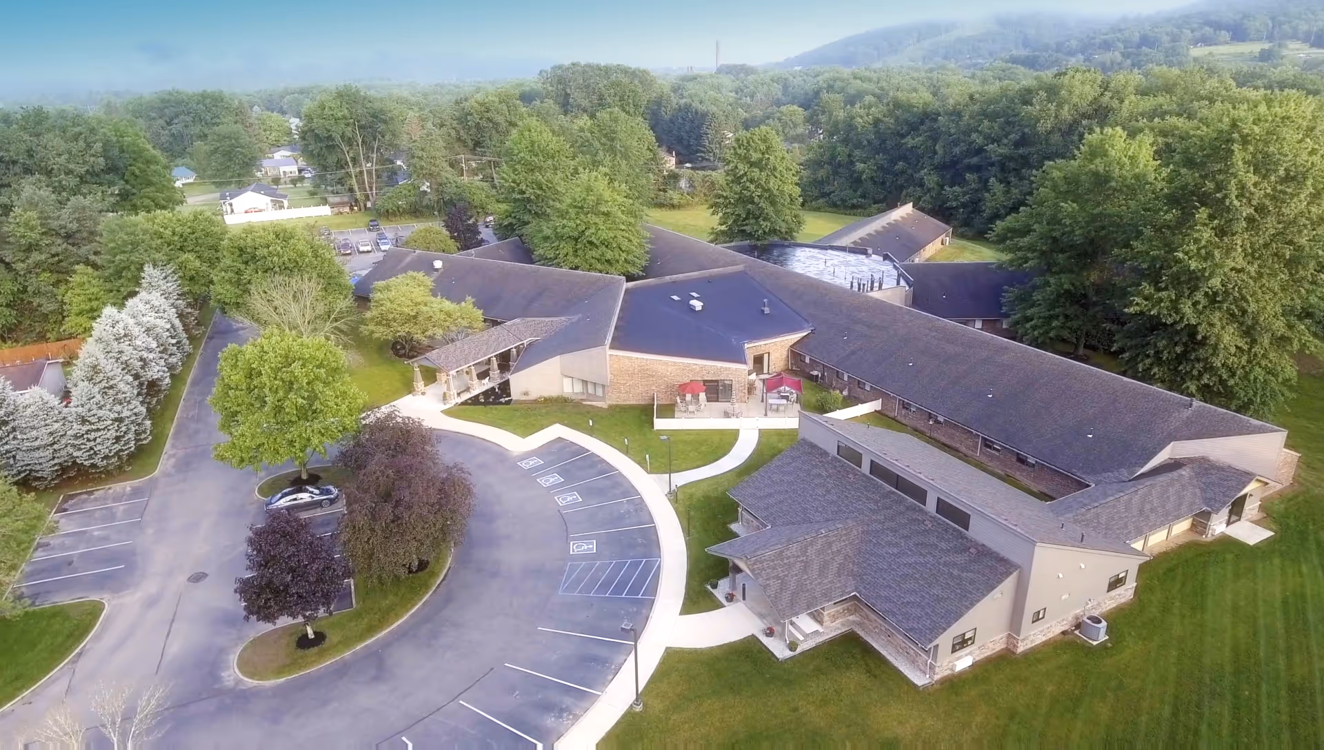 Aerial view of Warren Manor senior living facility surrounded by trees and greenery. The building has multiple wings with dark roofs and light-colored walls. There is a curved driveway with parking spaces, some marked for handicapped parking, and landscaped areas with trees and grass.