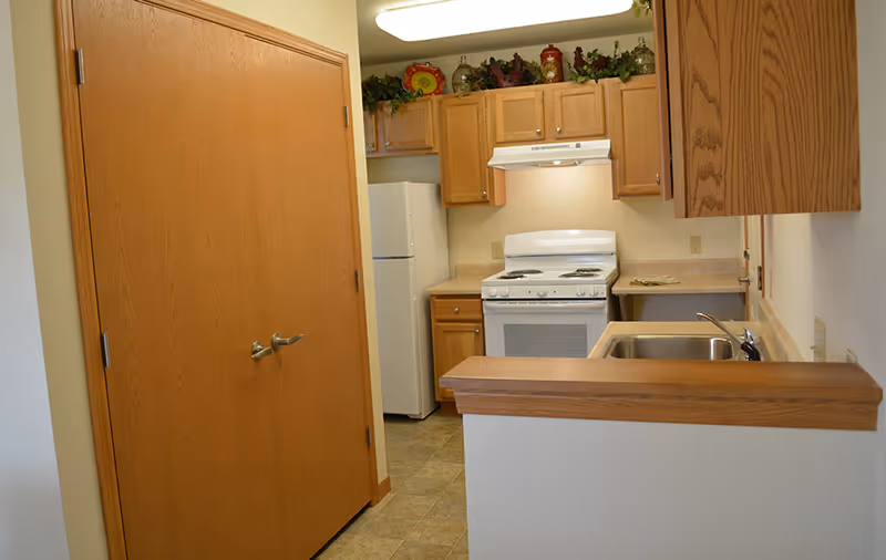 Small kitchen area with wooden cabinets, a white refrigerator, a white stove with an overhead vent, a sink with a faucet, and a wooden counter. There are decorative items on top of the cabinets and a double wooden door on the left side.