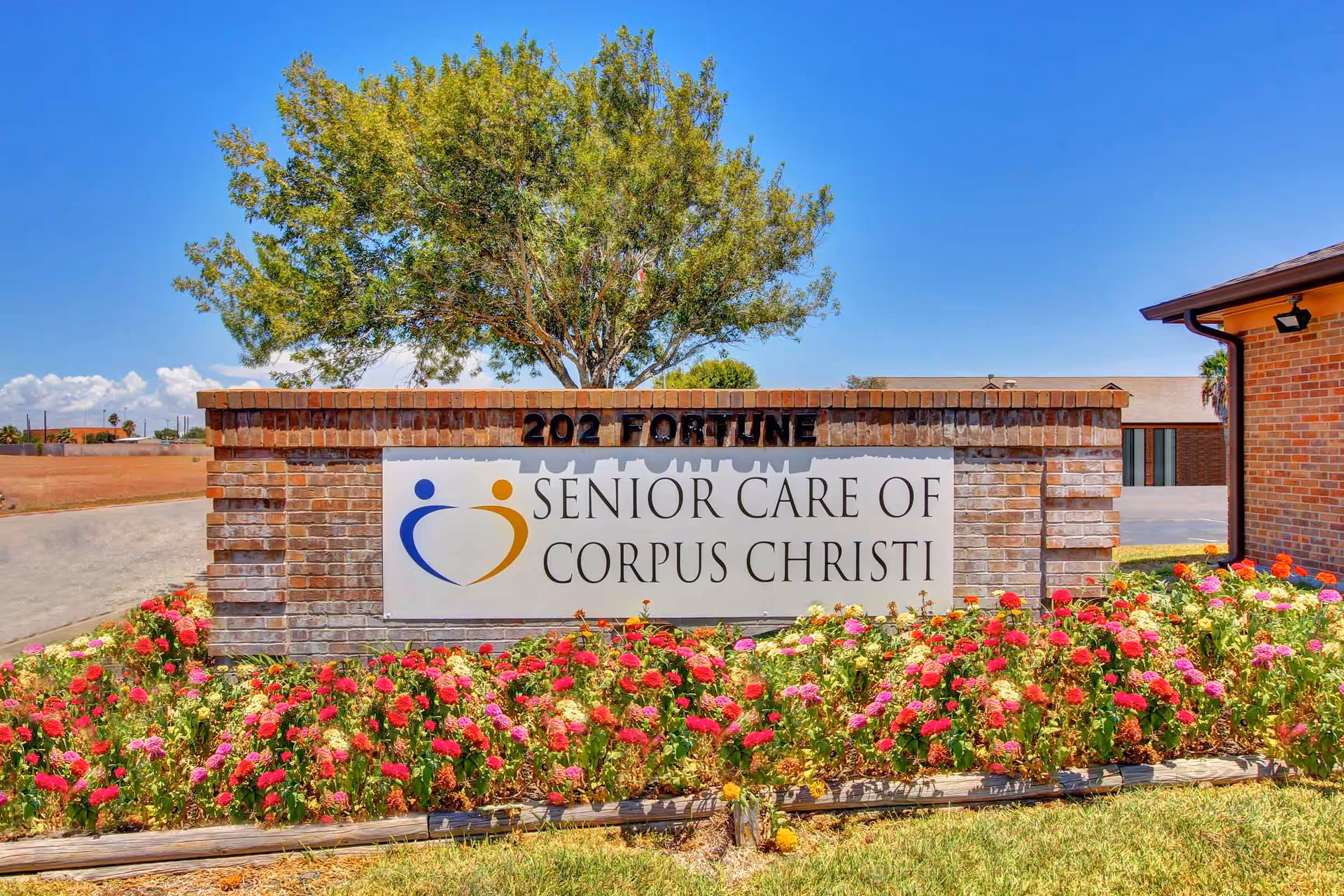 Brick sign with the text 'Senior Care of Corpus Christi' in front of a landscaped area with colorful flowers and a tree behind it under a clear blue sky.