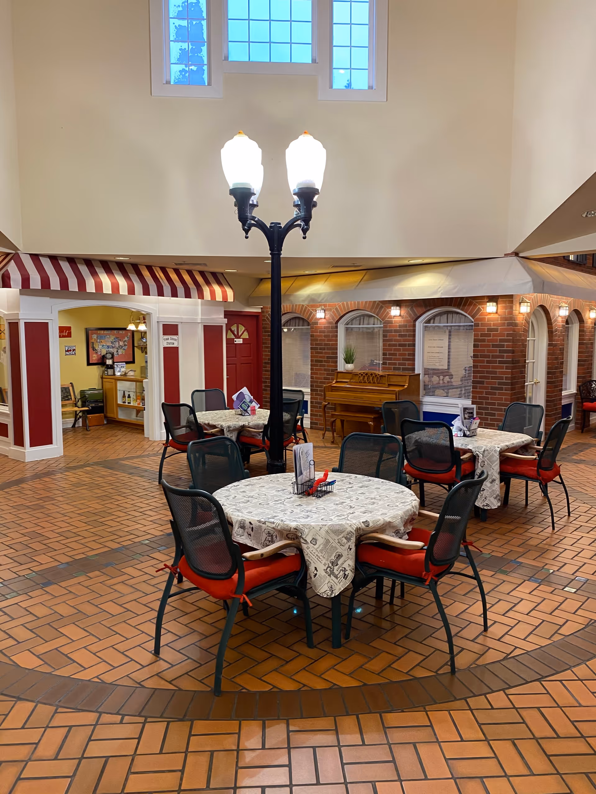 Indoor communal area with round tables covered in patterned tablecloths and surrounded by chairs with red cushions. A black streetlamp-style light fixture stands in the center. The background features a brick wall with windows, a piano, and a small storefront-style entrance with a red and white striped awning. Large windows near the ceiling allow natural light to enter.