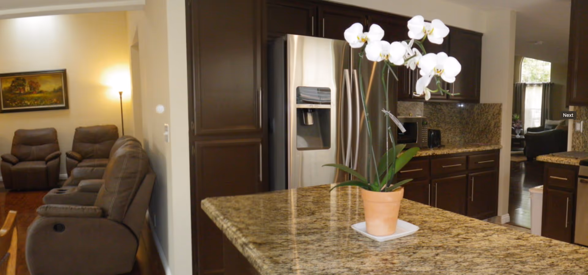 Interior view of a senior living facility showing a kitchen with granite countertops, dark wood cabinets, a stainless steel refrigerator, microwave, and a potted white orchid on the kitchen island. In the background, there is a living area with brown recliner chairs, a floor lamp, and a framed painting on the wall.