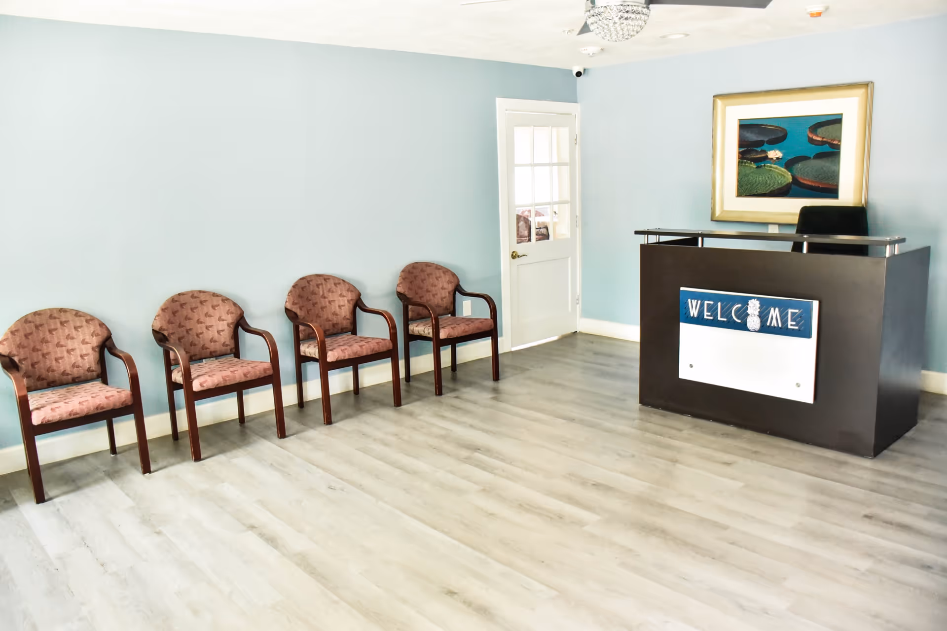 A waiting area with four wooden chairs with patterned upholstery lined up against a light blue wall. To the right, there is a dark reception desk with a 'WELCOME' sign featuring a pineapple design. Behind the desk is a black chair and a framed picture of lily pads on the wall. The floor is light-colored wood, and there is a white door with glass panels in the background.