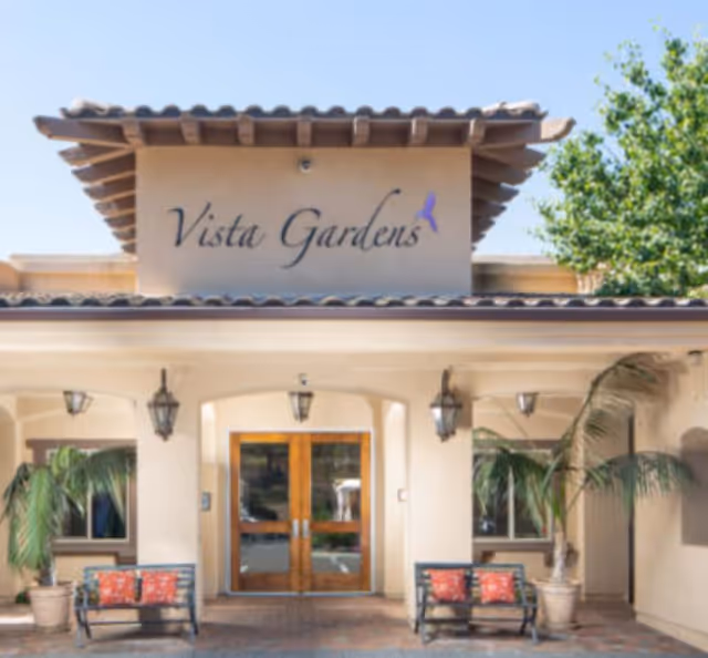 Front entrance of Vista Gardens with double glass doors, benches, potted palms and the 'Vista Gardens' sign above.