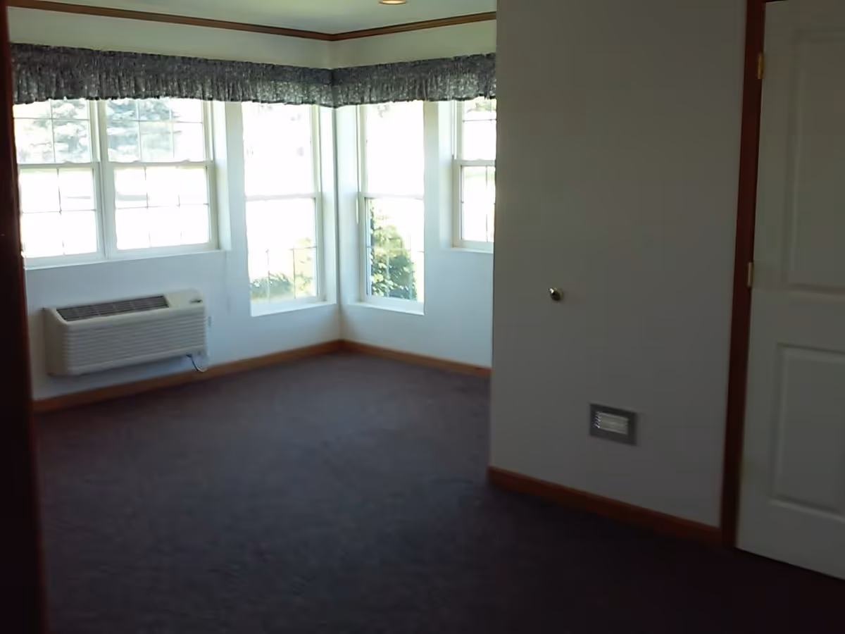 Empty carpeted room with bay windows, a wall air-conditioning unit, and a closed door.