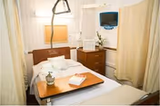 A hospital-style patient room with a single bed featuring white linens and pillows. There is a wooden overbed table with a small potted plant and a book on it. The room has beige curtains for privacy, a wooden bedside cabinet with a flower vase, and a wall-mounted television. Medical equipment is visible near the bed.