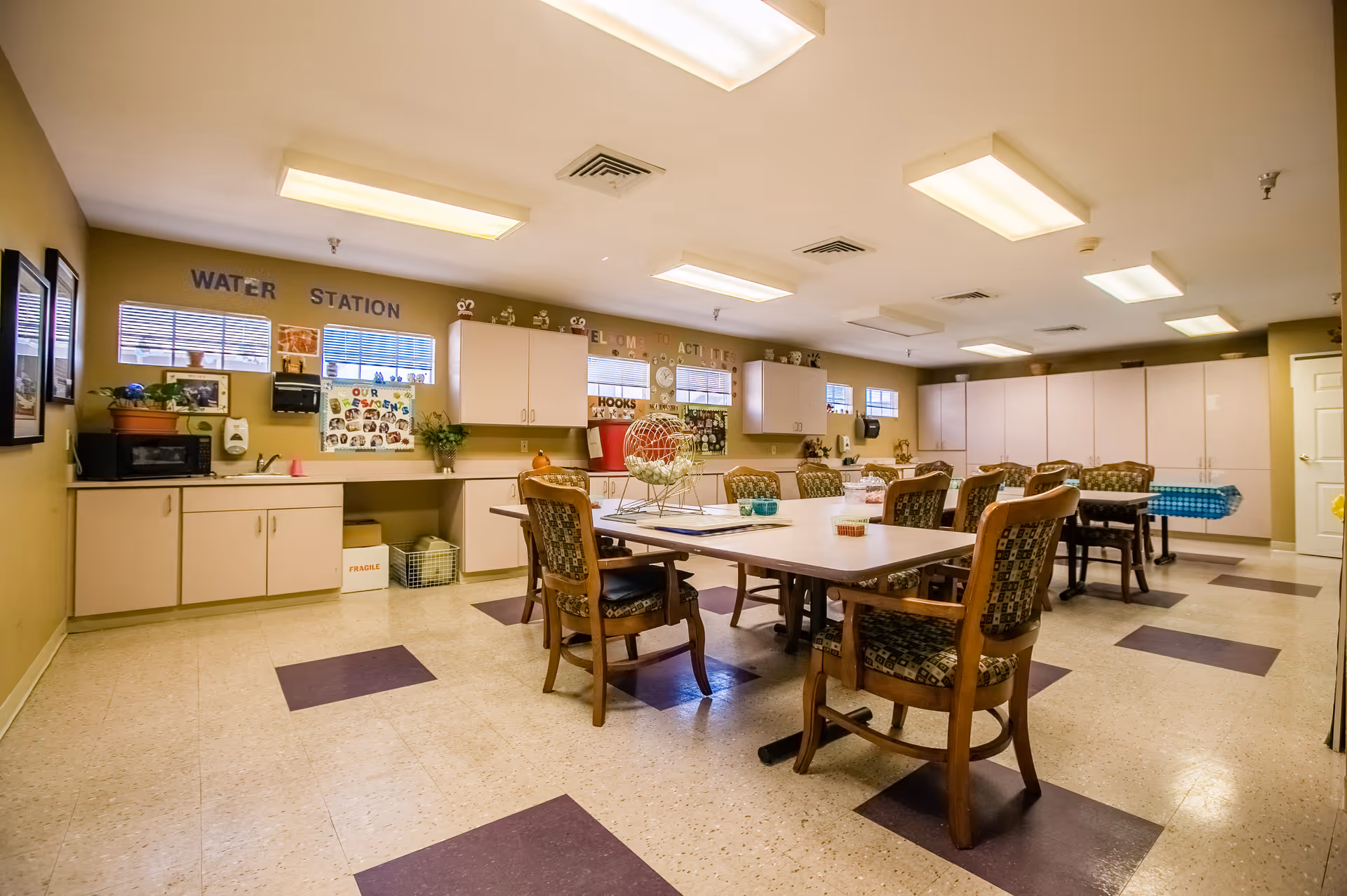 A well-lit activity room with tables and chairs arranged for group activities. The room has cabinets along the walls, a microwave, a sink, and various decorations including plants and posters. The floor has a pattern of beige and purple tiles, and fluorescent lights illuminate the ceiling.
