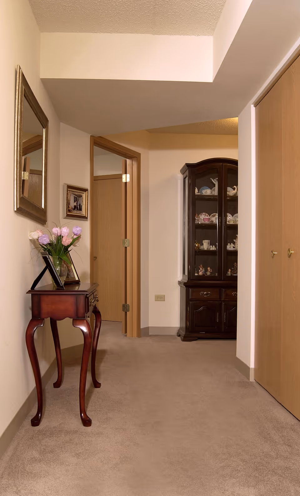 A hallway in a senior living facility with beige carpet and walls. On the left side, there is a small wooden table with curved legs holding a vase of pink and white flowers and a framed photo. Above the table is a large rectangular mirror and a small framed picture. At the end of the hallway, there is a wooden cabinet with glass doors displaying various decorative items and dishes. There are two wooden doors on the right side of the hallway.