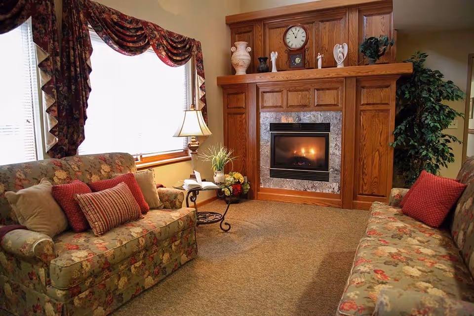 A cozy living room with two floral patterned sofas facing each other, a wooden fireplace with a clock and decorative items on the mantel, a side table with a lamp and an open book, and a large window with floral curtains letting in natural light.