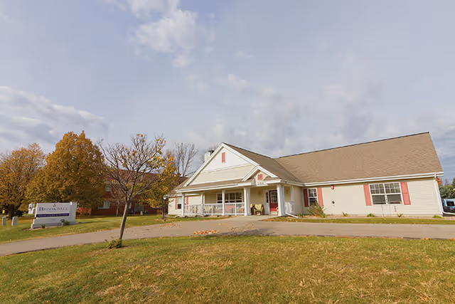 Single-story assisted living building with a covered front entrance, driveway and lawn under a cloudy sky.