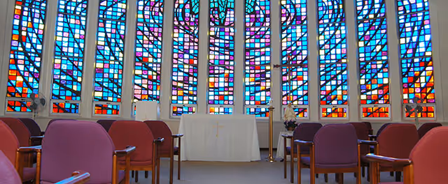 Interior view of a chapel or worship room with rows of purple and red chairs facing an altar covered with a white cloth. Behind the altar are large colorful stained glass windows with geometric patterns.