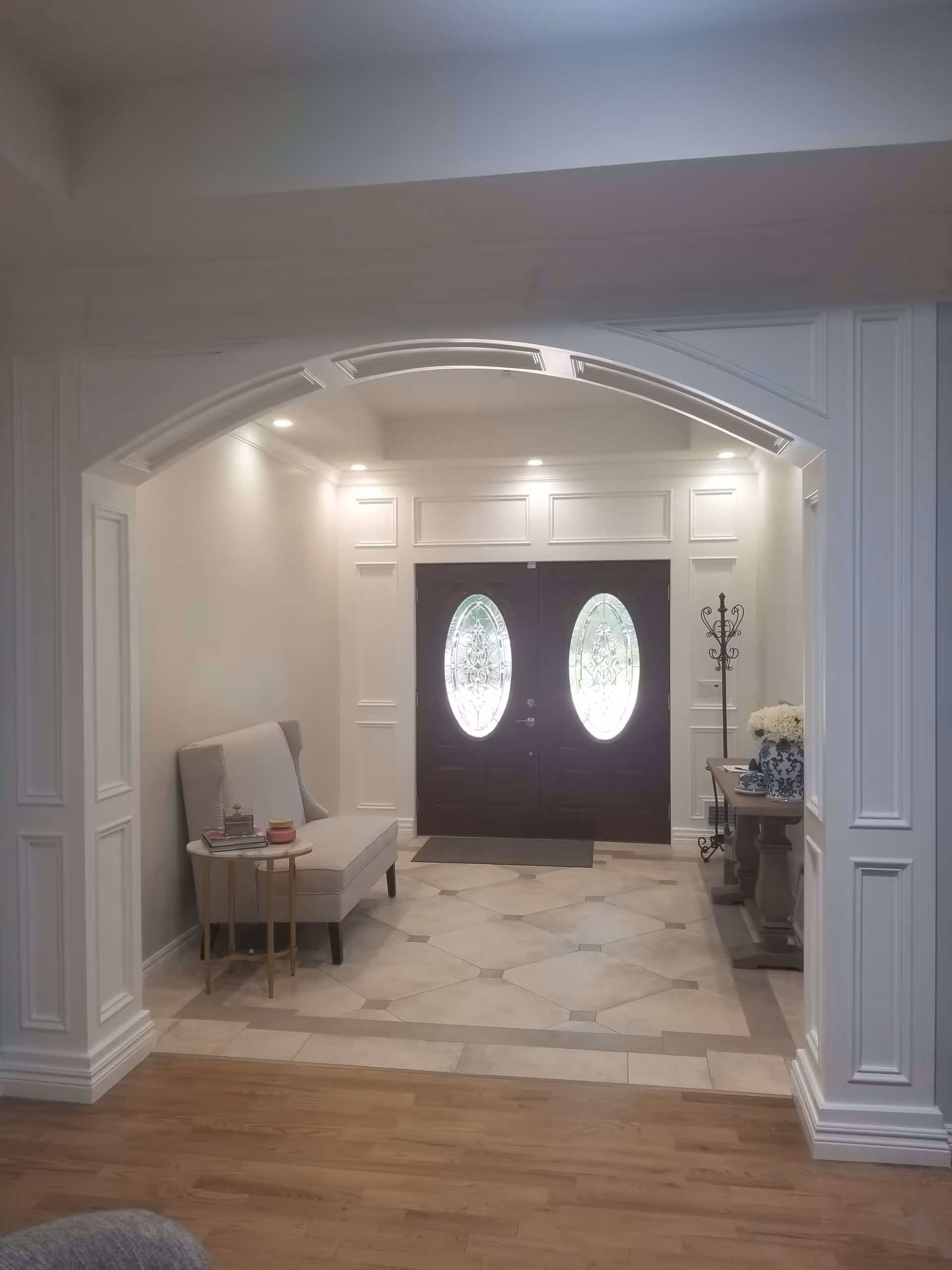 Interior view of an entryway with a double door featuring oval glass panels. The space has white walls with decorative molding, recessed ceiling lights, and a tiled floor. There is a beige upholstered bench with a small round side table on the left, and a wooden console table with a blue and white vase of flowers and a coat rack on the right.