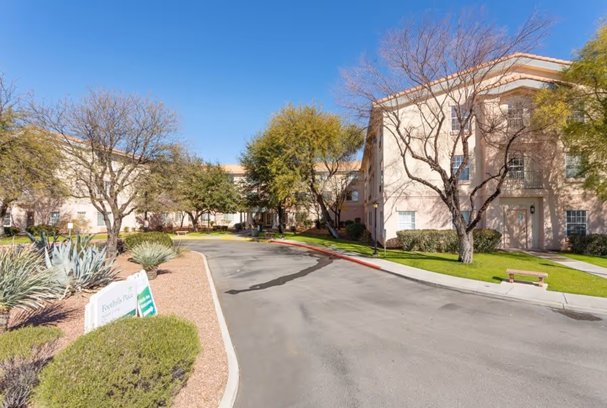 Exterior view of a senior living facility with a paved driveway, landscaped bushes, trees, and a clear blue sky. The building is light-colored with multiple windows and a bench near the sidewalk.