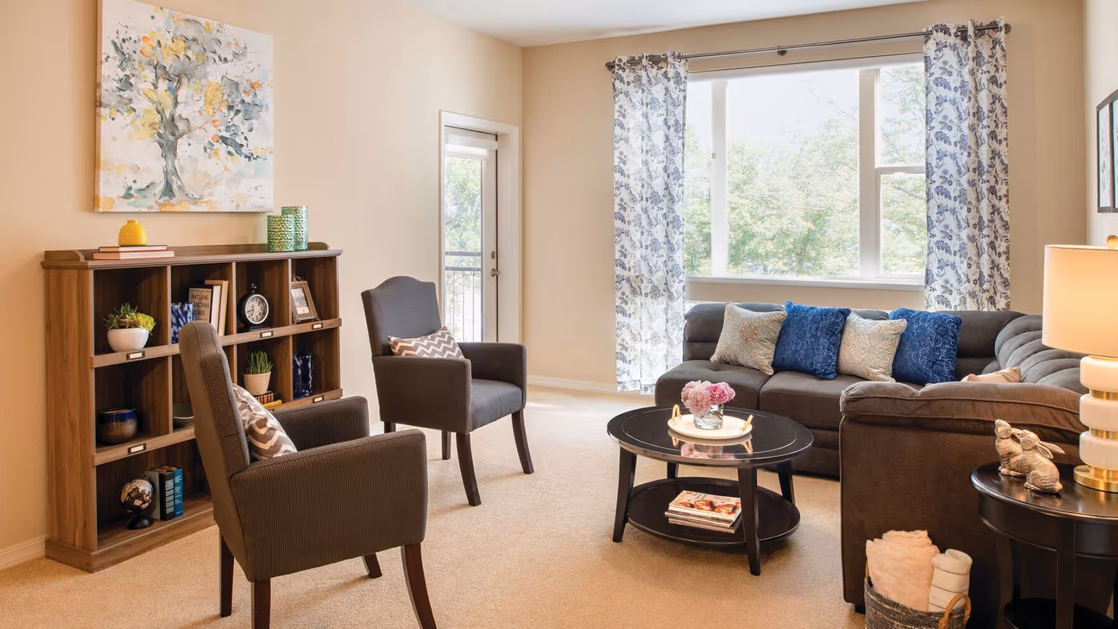 A bright and cozy living room with a large window covered by floral curtains, a gray sectional sofa with blue and beige pillows, two gray armchairs with patterned cushions, a round black coffee table with a flower vase and books underneath, a wooden bookshelf with decorative items and books, and a side table with a lamp and small sculptures.