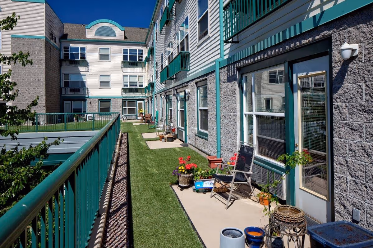 Outdoor patio area of Hazelwood Community Apartments with green railings, artificial grass, potted plants, chairs, and a multi-story building with windows and balconies under a clear blue sky.