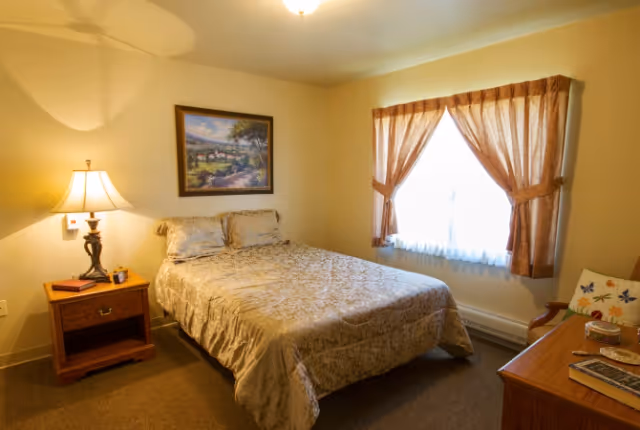 A simple furnished bedroom with a bed, nightstand and lamp, a window with curtains, and a dresser.