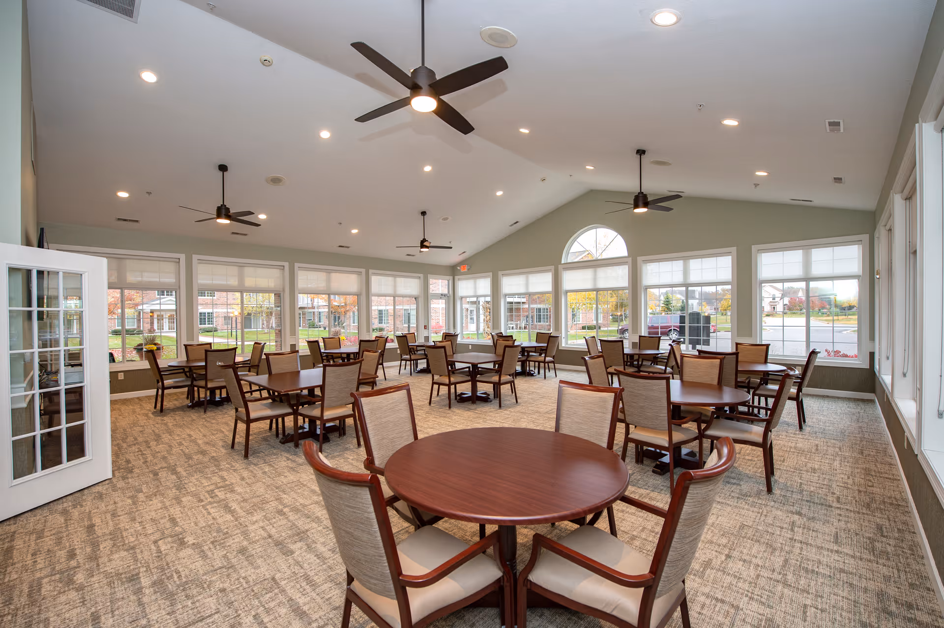 Bright communal dining room with multiple round and rectangular tables and chairs, large windows and ceiling fans.