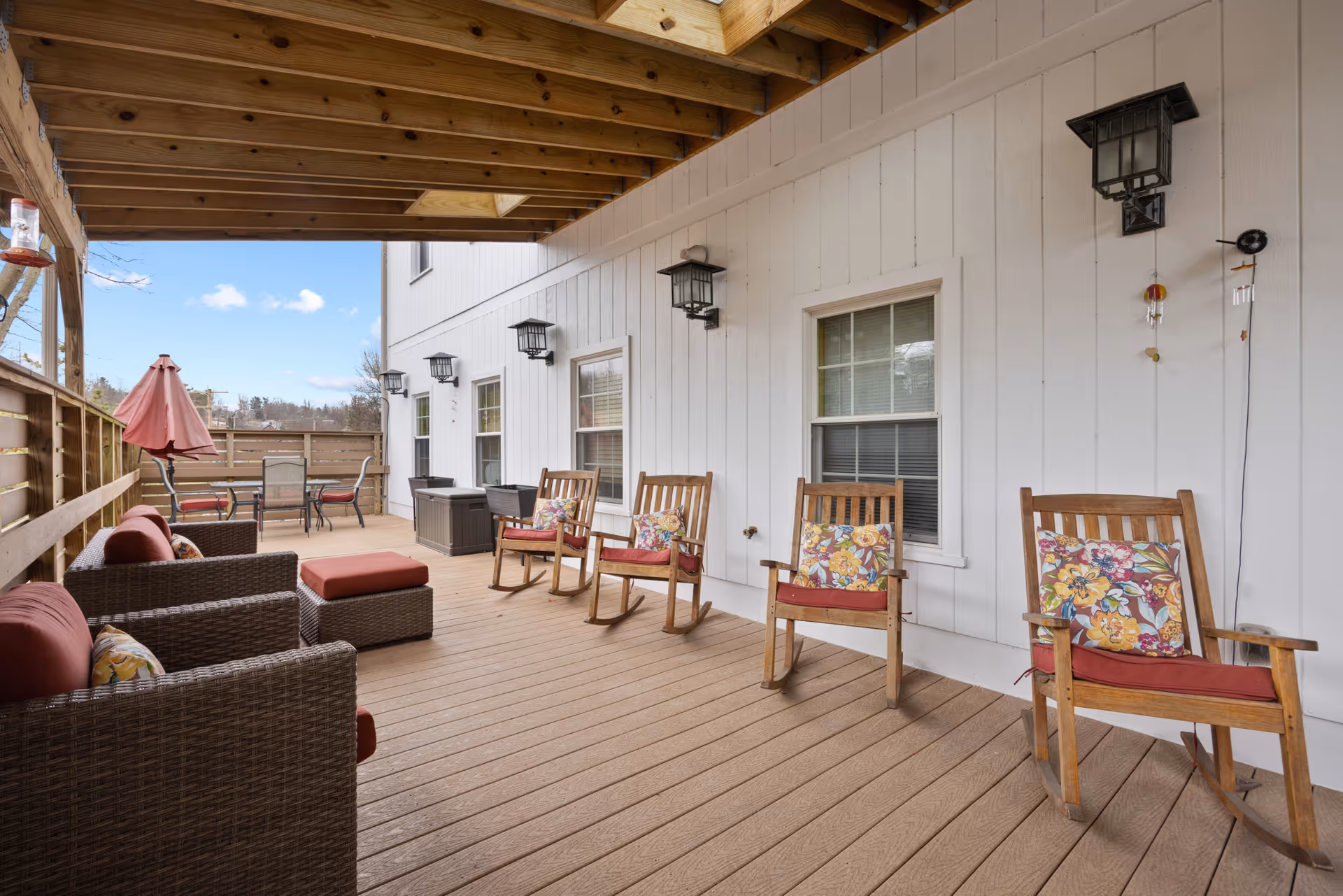 Covered outdoor patio area with wooden rocking chairs featuring floral cushions, wicker seating with red cushions, a small table, and a dining table with chairs and a red umbrella. The patio has a wooden floor and white siding walls with several black wall-mounted lanterns.