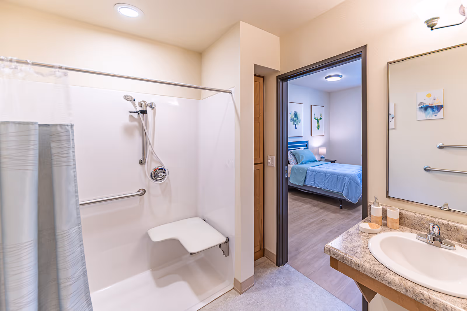 A clean and accessible bathroom with a shower area featuring a fold-down seat and grab bars. The shower has a handheld showerhead and a curtain. To the right, there is a sink with a countertop, soap dispenser, and a mirror above it. Through the doorway, a bedroom with a bed, nightstand, lamp, and framed artwork on the walls is visible.