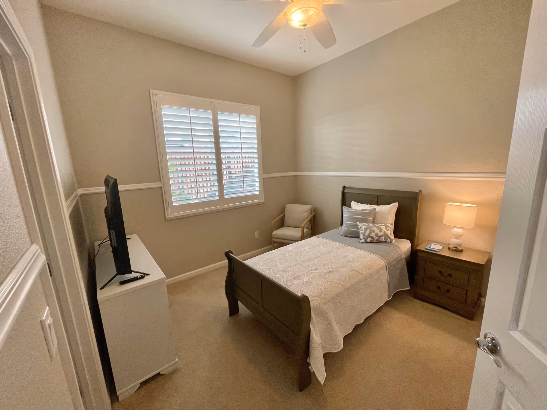 Neat single bedroom with a twin bed, nightstand and lamp, TV on a dresser, a chair, and a window with shutters.