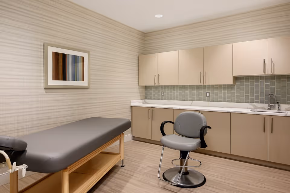 Clinical exam room with an upholstered examination table, a swivel chair, and beige cabinets with a sink and countertop.