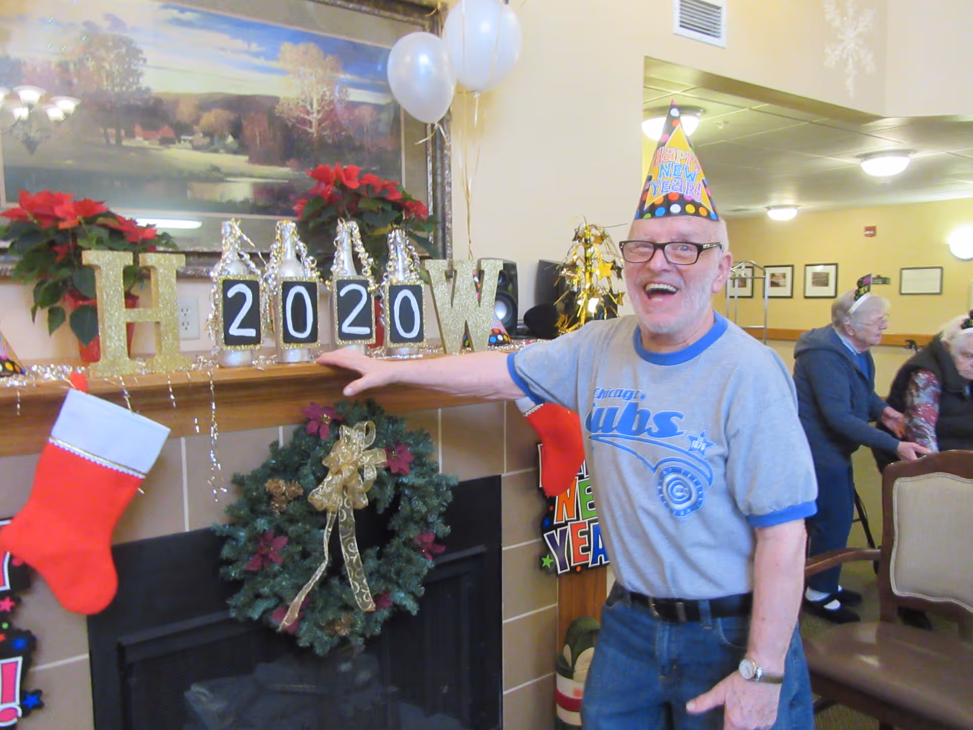 An elderly man wearing a 'Happy New Year' party hat and a Chicago Cubs t-shirt stands smiling next to a decorated fireplace mantel. The mantel is adorned with gold glitter letters spelling 'H 2020 W', red poinsettias, white balloons, and a Christmas wreath hanging below. In the background, two elderly women are seated and engaged in conversation in a well-lit room.