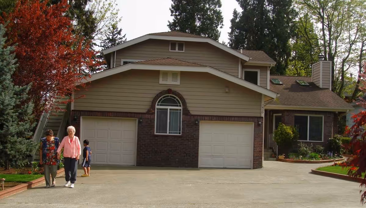 Front exterior of a two-story suburban house with a double garage, driveway, landscaping, and three people walking near the entrance.