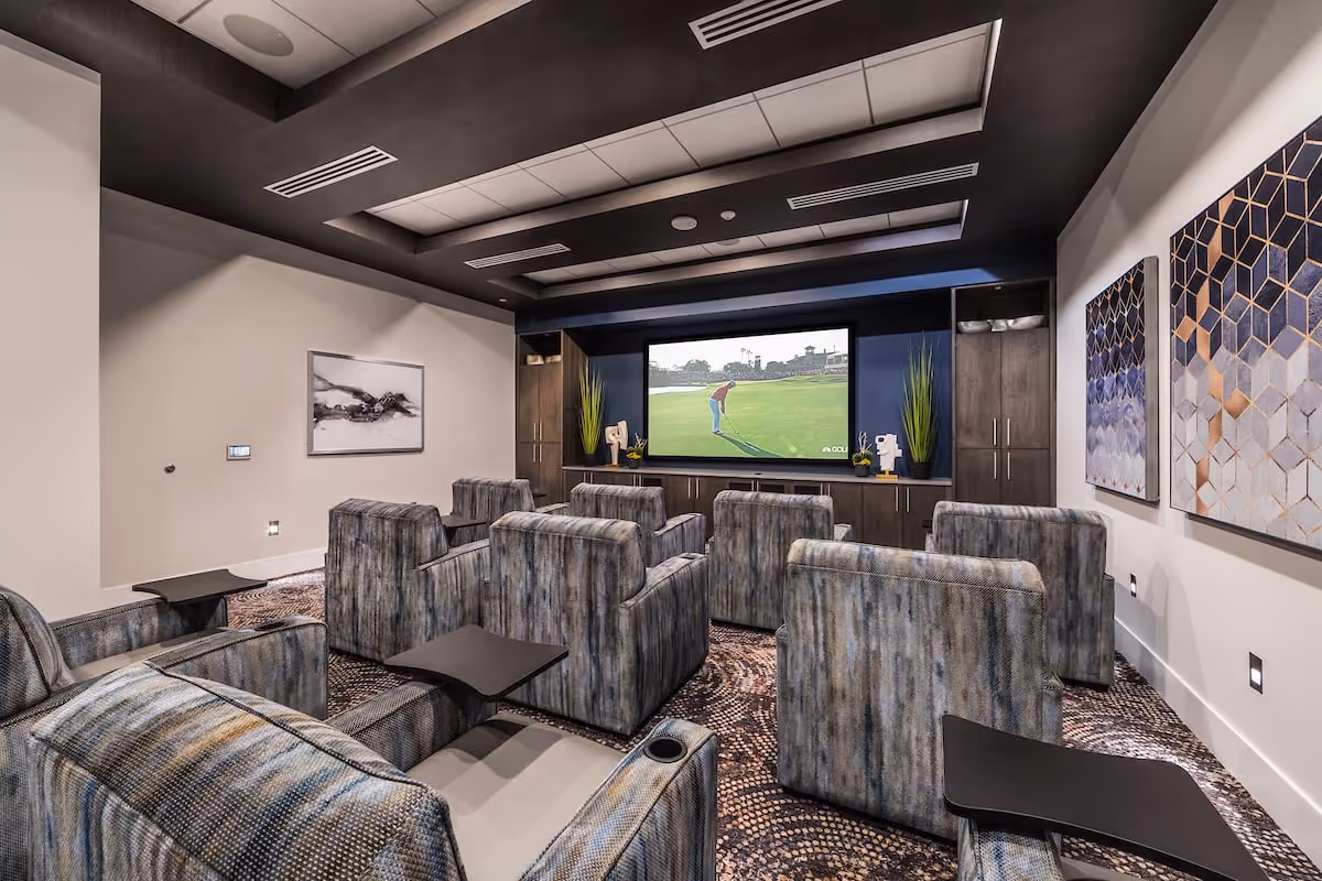 A modern media room with multiple comfortable upholstered armchairs arranged in rows facing a large flat-screen TV mounted on a dark wood entertainment center. The room features a patterned carpet, recessed lighting in the ceiling, abstract artwork on the walls, and decorative plants on the entertainment center.