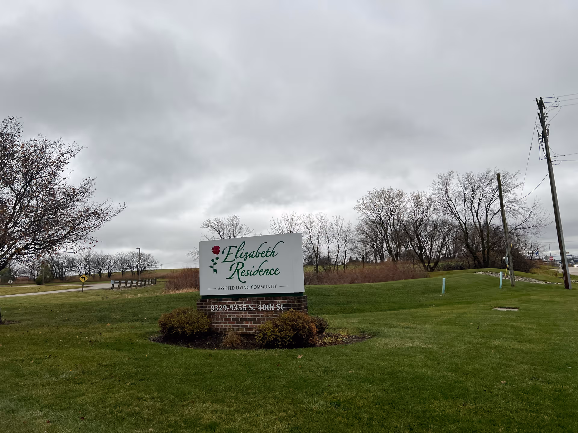 Outdoor view of a grassy area with a sign for Elizabeth Residence Assisted Living Community. The sign is white with green text and a red rose graphic, mounted on a brick base. Leafless trees and a cloudy sky are visible in the background.