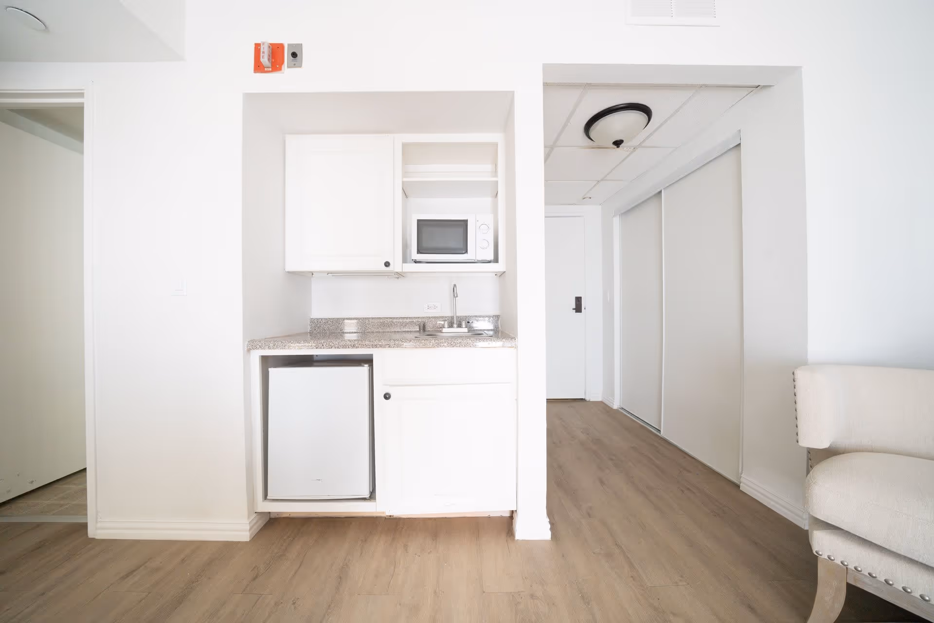 Small kitchenette area with white cabinets, a microwave, a mini refrigerator, and a sink. The floor is light wood, and there is a white door and closet with sliding doors in the background. Part of a beige upholstered chair is visible on the right side.