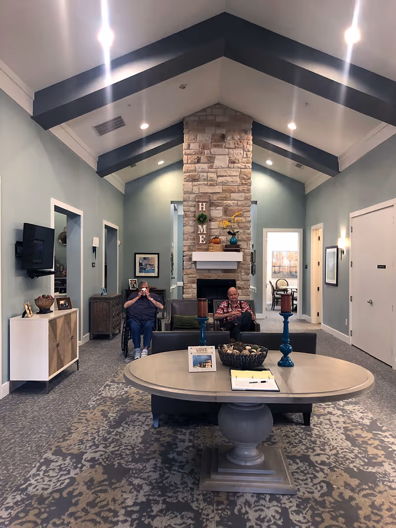 Interior of a senior living facility common area with a high vaulted ceiling featuring dark wooden beams. A stone fireplace is centered on the far wall with decorative items including a sign spelling HOME. Two elderly men are seated near the fireplace, one in a wheelchair drinking from a cup and the other sitting on a chair. A round table with candles and a basket is in the foreground, and there is a TV mounted on the left wall above a cabinet. The room has soft blue walls and patterned carpet.