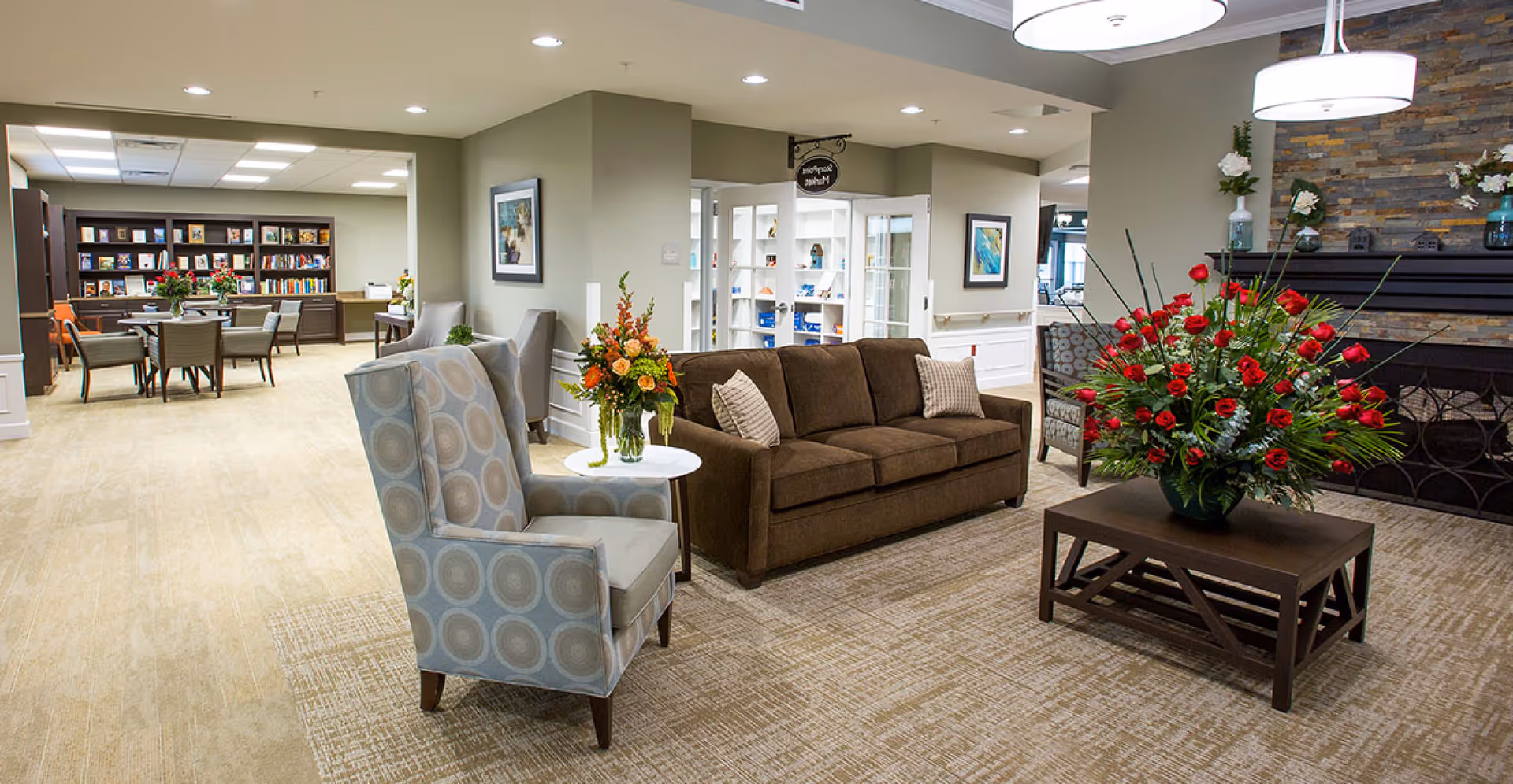Spacious senior living common area with sofas, armchairs, a coffee table topped by a large floral arrangement, and a reading area in the background.