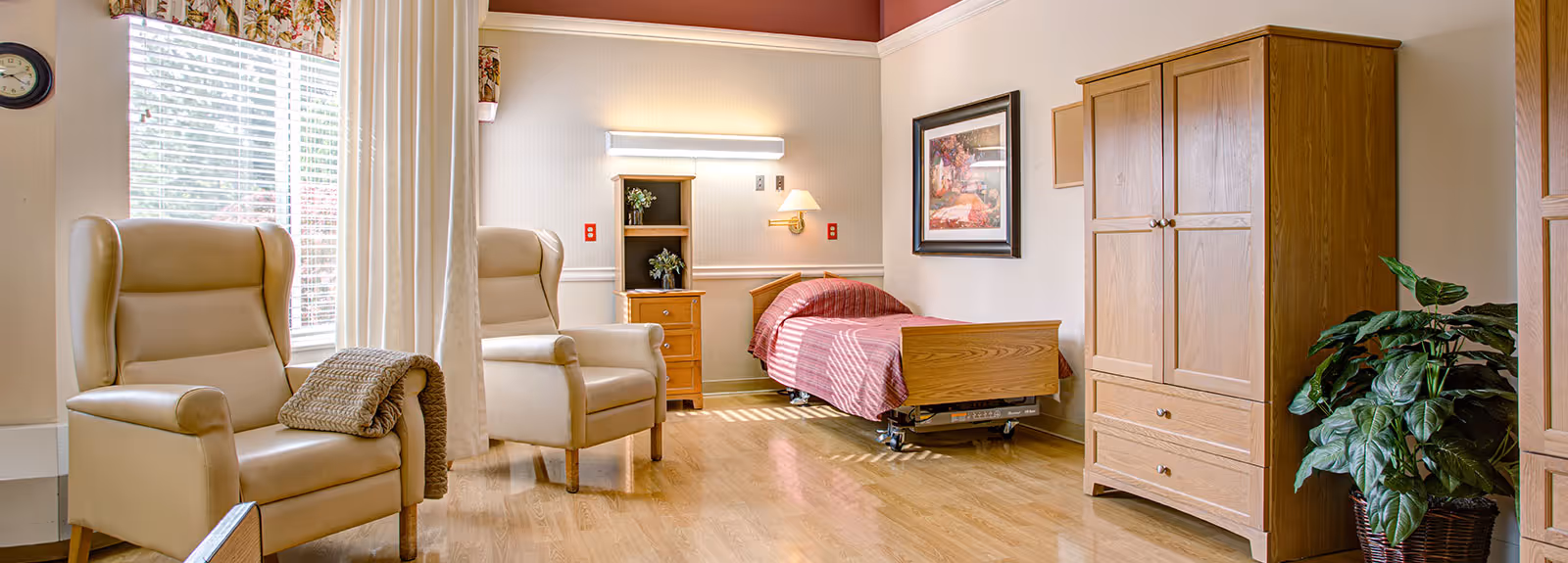 Sunny assisted-living bedroom with two beige armchairs, a single adjustable bed with red cover, wooden wardrobe and potted plant.