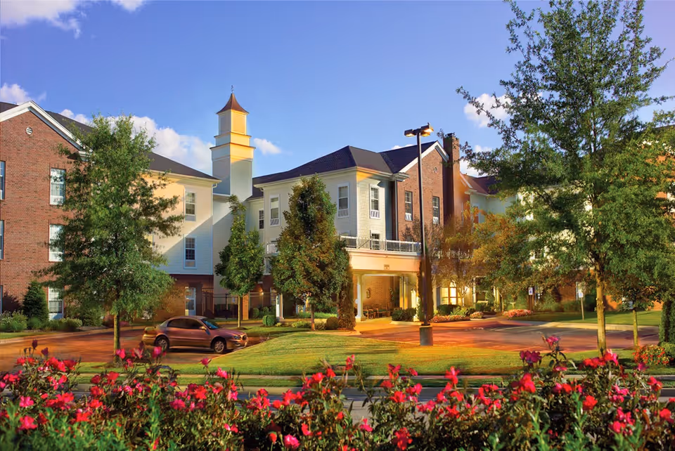 Exterior view of Belmont Village Senior Living St. Matthews building with a mix of brick and light-colored siding, surrounded by trees and vibrant red flowers in the foreground under a clear blue sky.