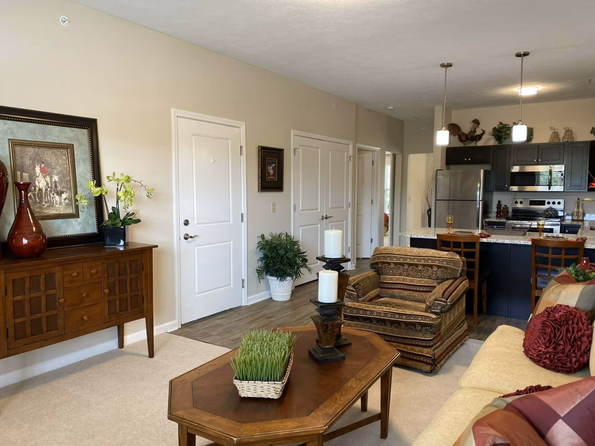 Open living room with sofa, armchair, coffee table and decorative plants, opening to a kitchen with island and bar stools.