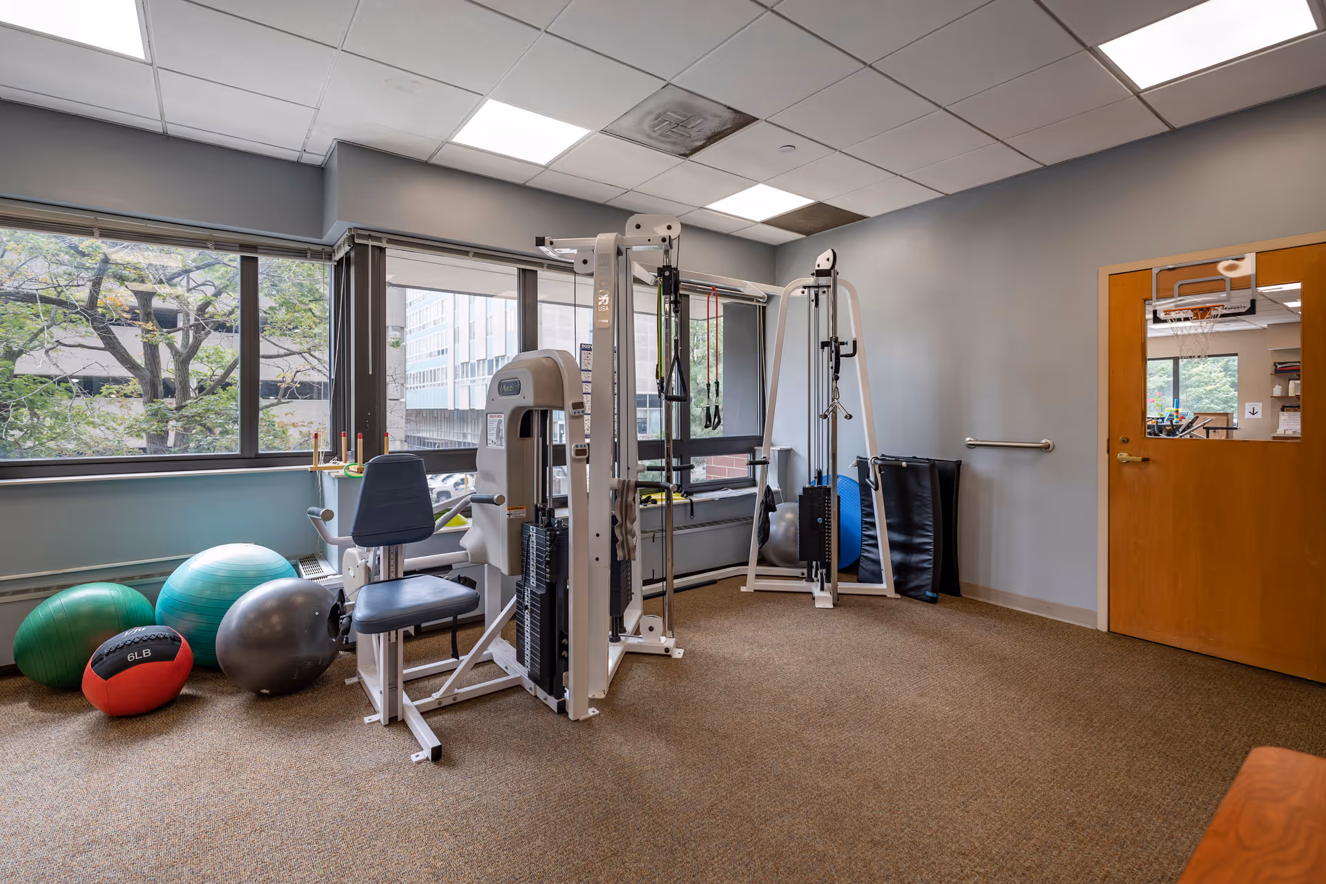 Small fitness room with weight machines, exercise balls, and large windows overlooking trees.