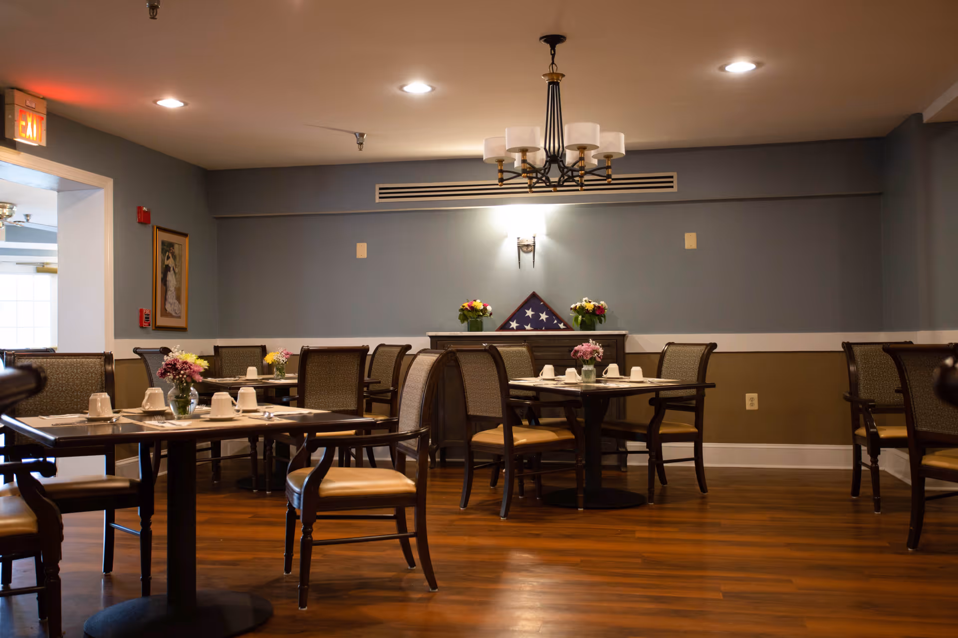 A dining room with several tables and chairs arranged neatly. Each table has white cups and small flower arrangements. The room has wooden flooring, a blue and beige wall with a folded American flag displayed on a sideboard, and a chandelier hanging from the ceiling. There is an exit sign visible above a doorway on the left side.