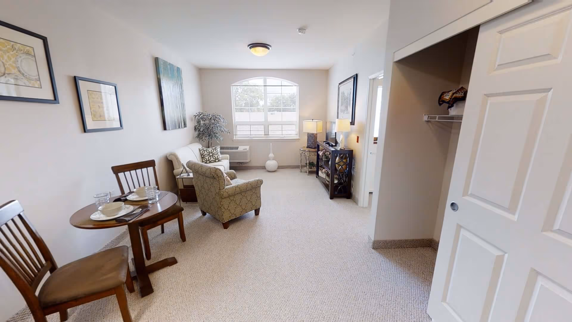 Bright furnished living room with a small dining table, armchairs, TV console, and a window letting in natural light.