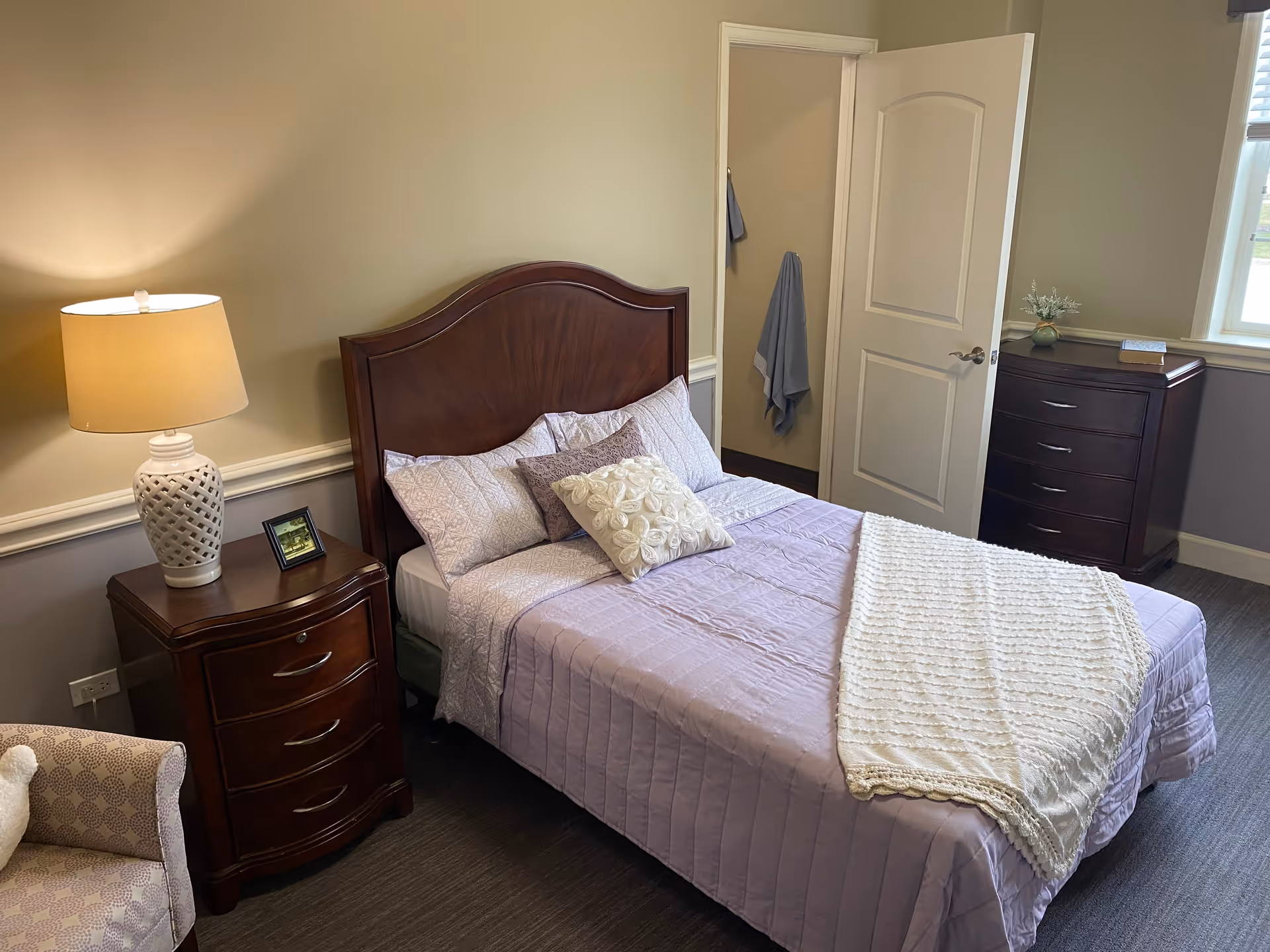 A neatly made bed with a wooden headboard in a bedroom. The bed has light purple bedding with decorative pillows and a white knitted throw blanket. To the left of the bed is a wooden nightstand with a white ceramic lamp and a small framed picture. There is a patterned armchair partially visible in the bottom left corner. An open door reveals a towel hanging inside a closet or bathroom. To the right, there is a wooden dresser with a small plant and a book on top, and a window with blinds letting in natural light.