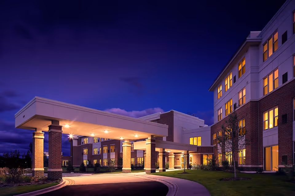 Exterior view of a multi-story senior living facility at dusk with warm lights glowing from the windows. The building features brick and light-colored siding with a covered entrance supported by brick columns. The sky is deep blue with some clouds, and the driveway curves under the covered entrance.