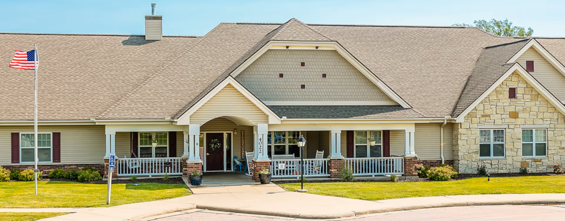 Front exterior view of a single-story senior living facility building with beige siding, stone accents, a covered entrance with white columns, an American flag on a flagpole, and a well-maintained lawn.