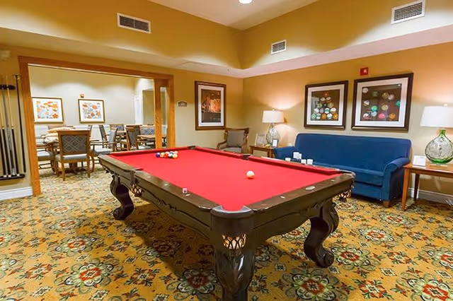 A recreational room with a red felt pool table in the center, surrounded by a patterned carpet. In the background, there is a blue couch with two framed artworks above it, a side table with a lamp and decorative items, and an adjacent room with a dining table and chairs. The walls are painted a warm beige color.