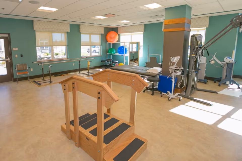 A bright physical therapy room with wooden practice stairs in the foreground, parallel bars, therapy table, exercise machines, and large stability balls by the windows.