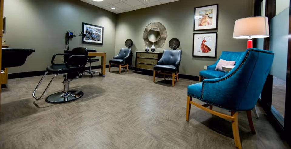 A modern salon-style seating area with teal armchairs, black styling chairs, a dresser, round mirror and framed wall art in a well-lit room.