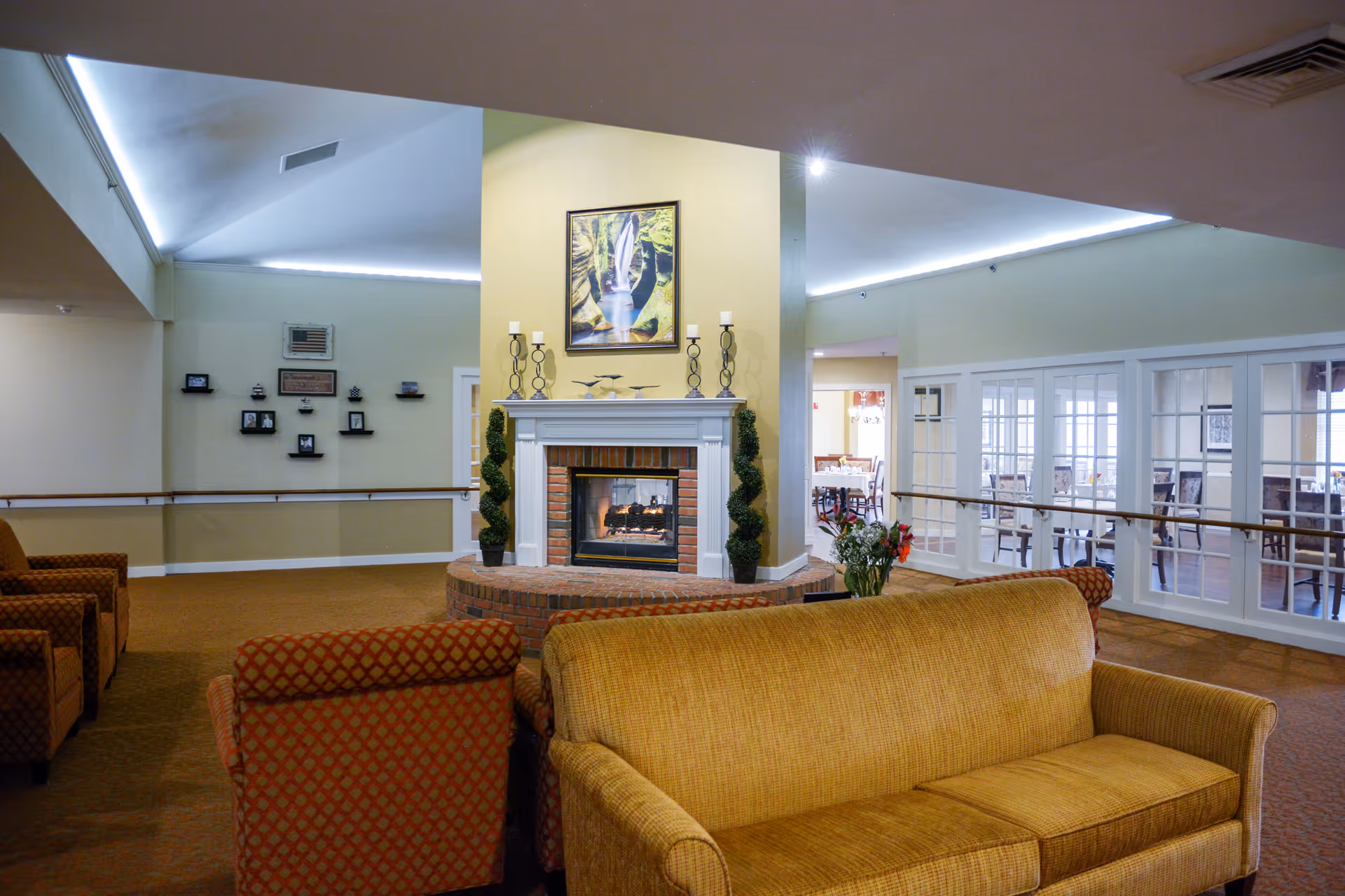 A cozy senior living facility common area with a central brick fireplace surrounded by a white mantel, decorated with candle holders and small topiary plants. The room features comfortable upholstered armchairs and a sofa in warm tones. In the background, a dining area with tables and chairs is visible through glass-paneled doors. The walls are painted light yellow and the ceiling has recessed lighting.
