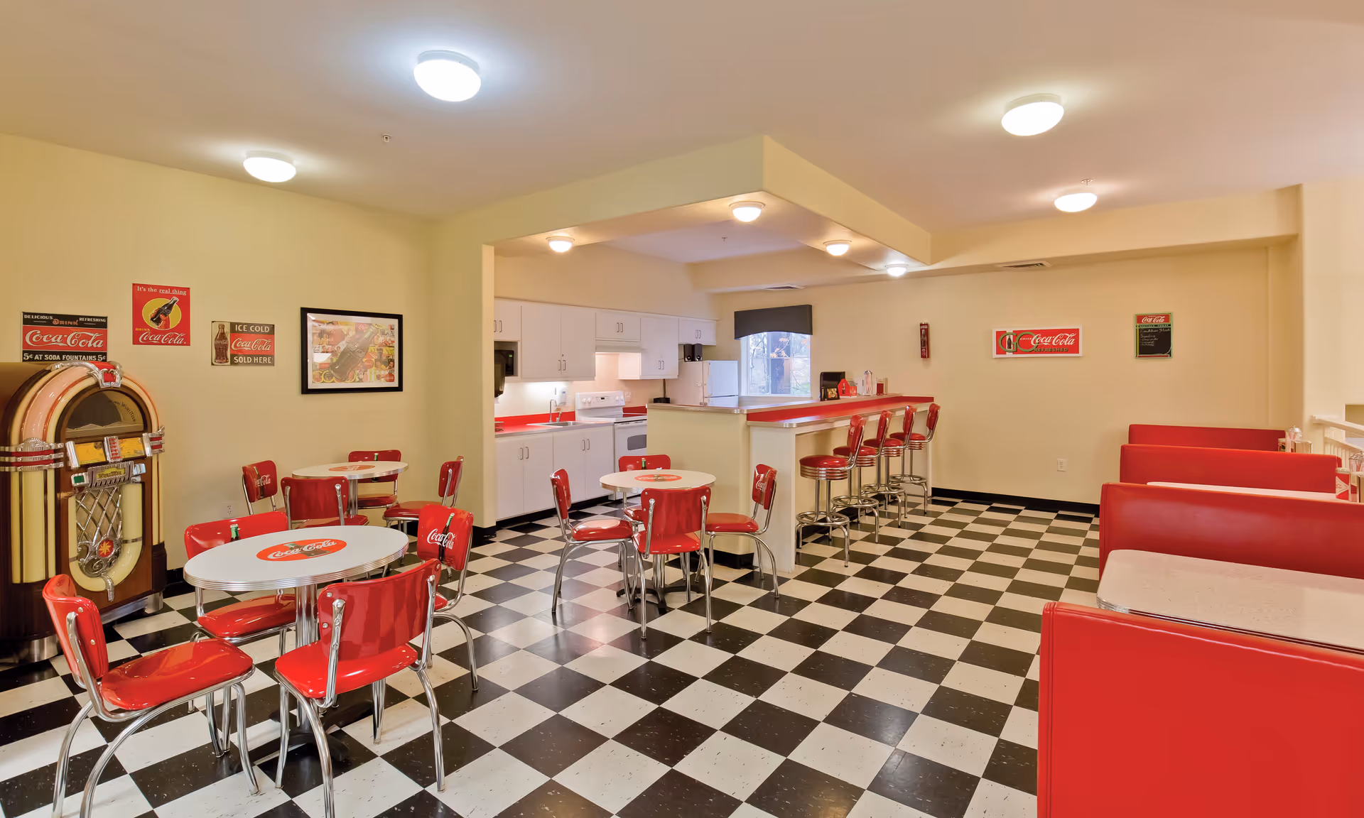 Retro-style dining room with red booths and chrome chairs around round tables, a checkered black-and-white floor, a jukebox, and a counter with barstools.