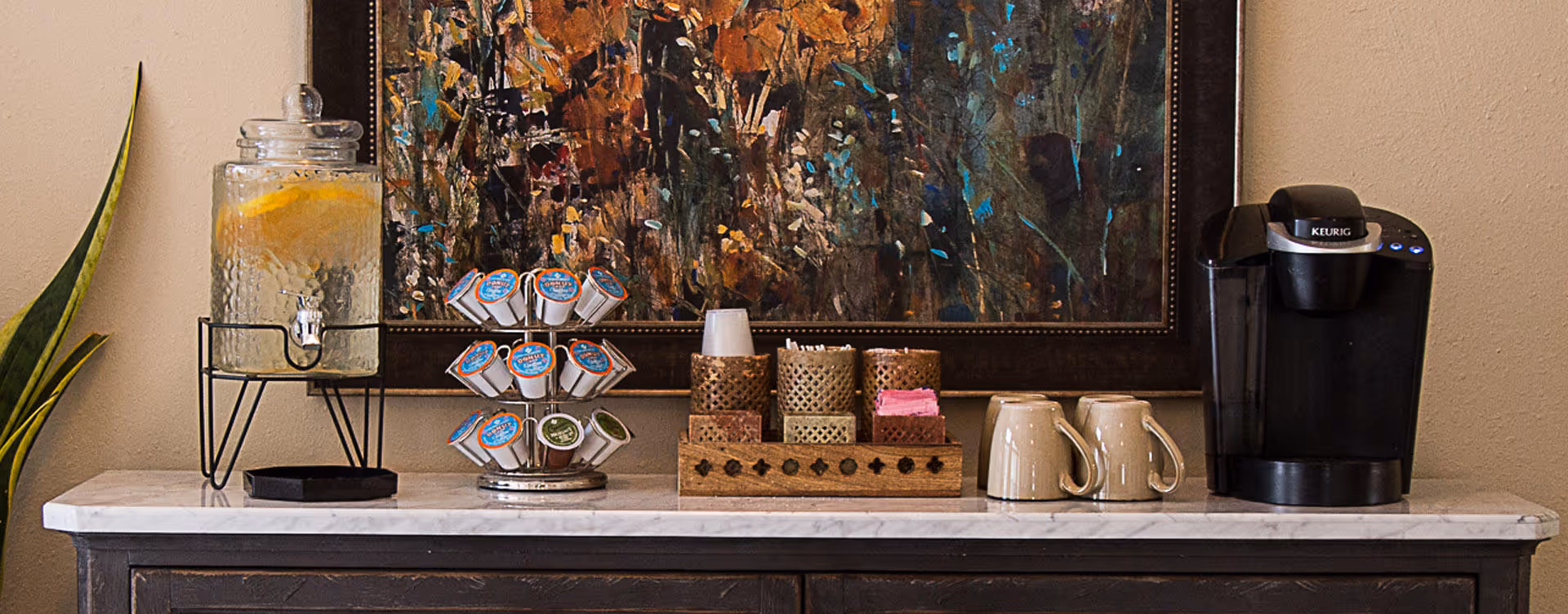 A countertop setup featuring a glass dispenser with lemon water, a rotating holder with coffee pods, a wooden tray holding cups, napkins, and stirrers, two beige mugs, and a black Keurig coffee maker. A colorful abstract painting is hung on the wall behind the countertop.
