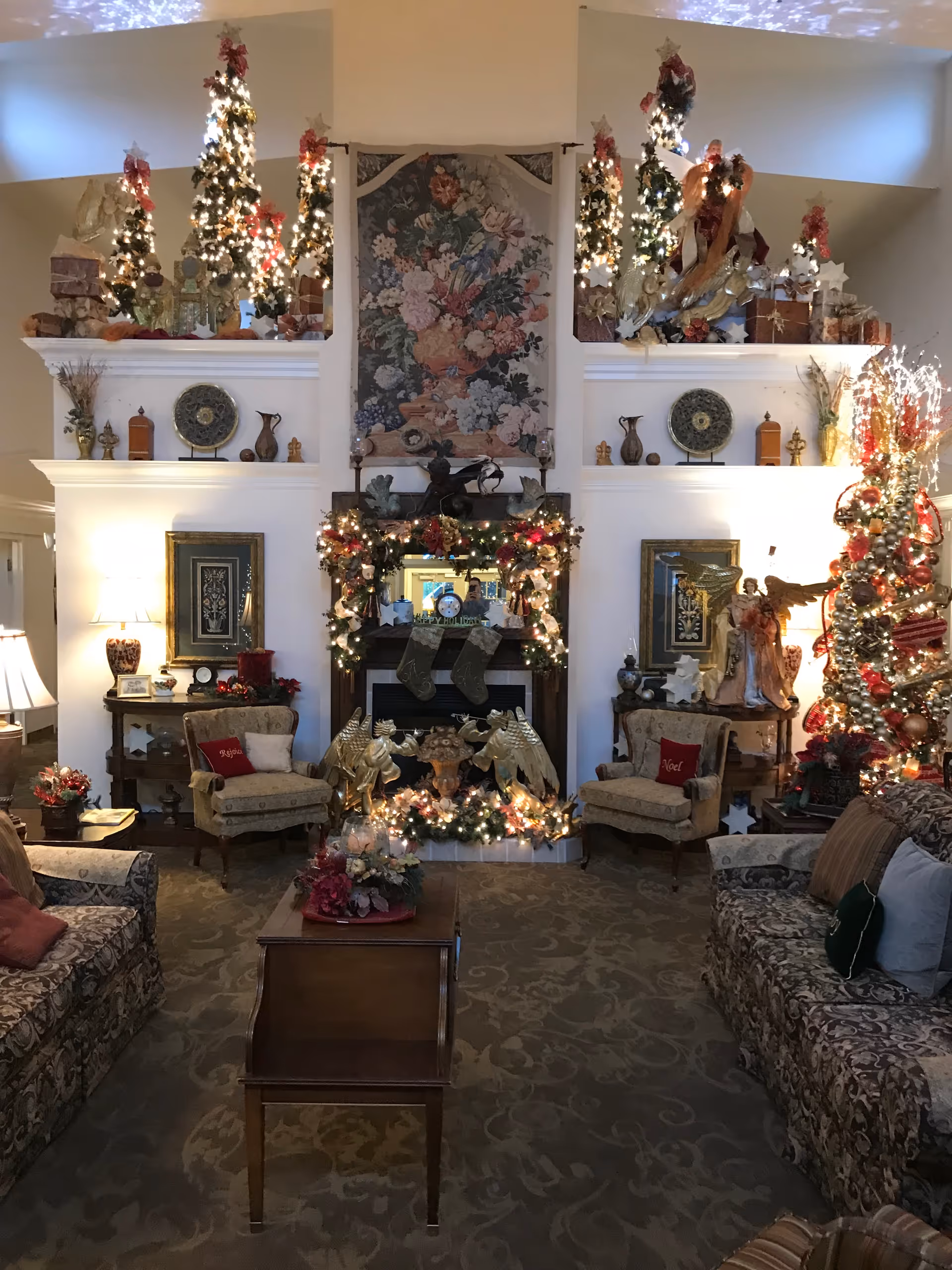 A warmly decorated living room with Christmas decorations including a lit Christmas tree, garlands with lights around a fireplace, and small decorated Christmas trees on shelves. The room has patterned sofas and armchairs, a wooden coffee table, framed artwork, and various decorative items. The lighting is soft and cozy, creating a festive atmosphere.