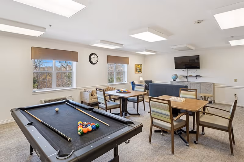 A well-lit recreational room with a black pool table in the foreground, two wooden tables with chairs, a blue couch, a wall-mounted TV, a globe on a cabinet, and two windows with brown valances letting in natural light.