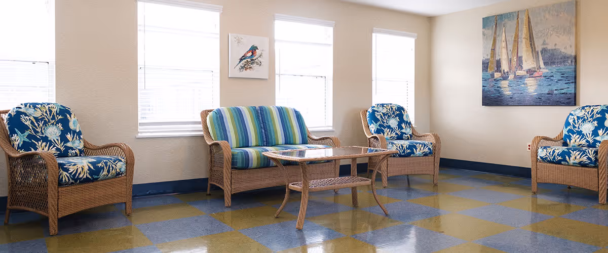 A bright sitting area with three wicker armchairs and one wicker loveseat, all with colorful cushions. The armchairs have blue cushions with white coral patterns, and the loveseat has striped cushions in shades of blue and green. A wicker coffee table with a glass top is centered in front of the seating. The room has three windows with white blinds, beige walls, and two paintings on the walls, one depicting a bird and the other sailboats on water. The floor has a checkered pattern in blue and green tiles.