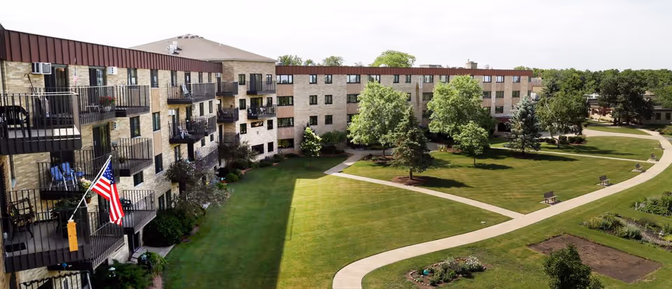 Exterior view of a multi-story senior living facility named Cedar Crest with balconies, an American flag, well-maintained green lawns, trees, walking paths, and benches in the garden area.